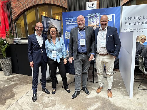 Vier Personen in formeller Kleidung stehen vor einem Stand eines Branchenevents. 