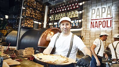 A pizza chef holds a freshly prepared pizza up to the camera