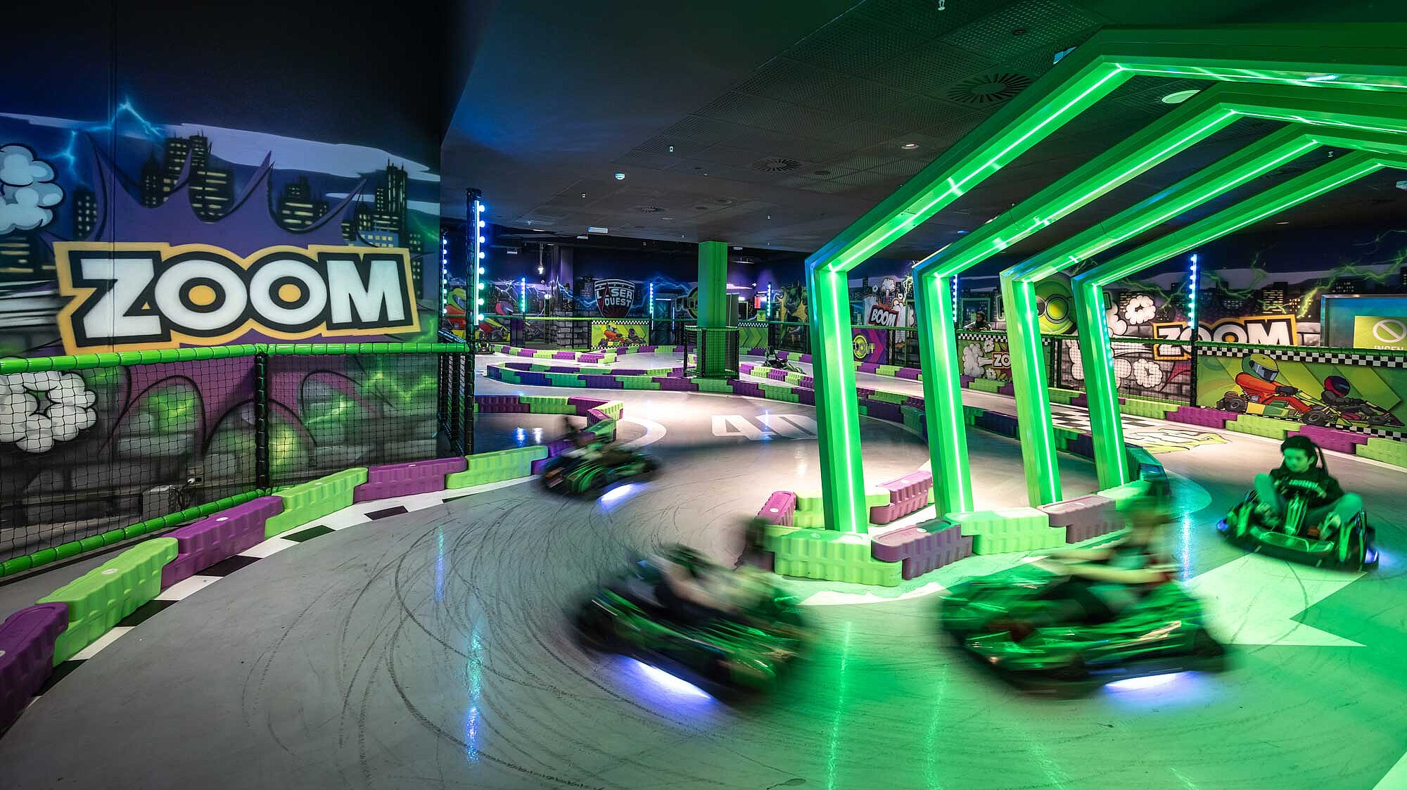 Es ist eine Kart-Bahn zu sehen, mit grüner Beleuchtung, wo vier Kinder entlang fahren.