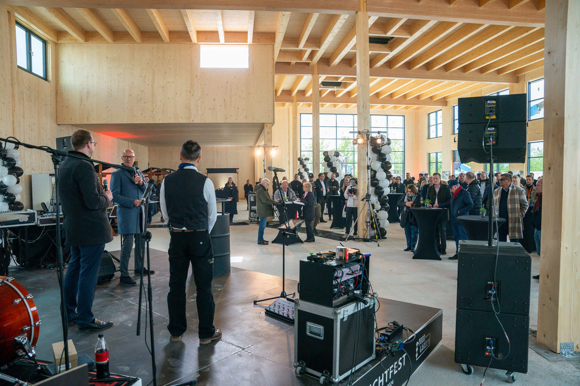 Drei Personen mit Mikrofonen auf Bühne, im festlich geschmückten Raum steht Publikum im Hintergrund.